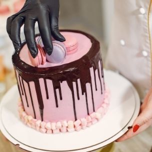 female baker in uniform and latex gloves decorating cake with macarons in light kitchen